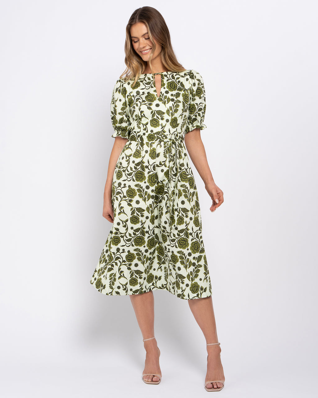 A woman modeling a knee-length midi dress with a green and off-white floral print, featuring puff sleeves and a keyhole neckline, set against a plain light gray studio background.