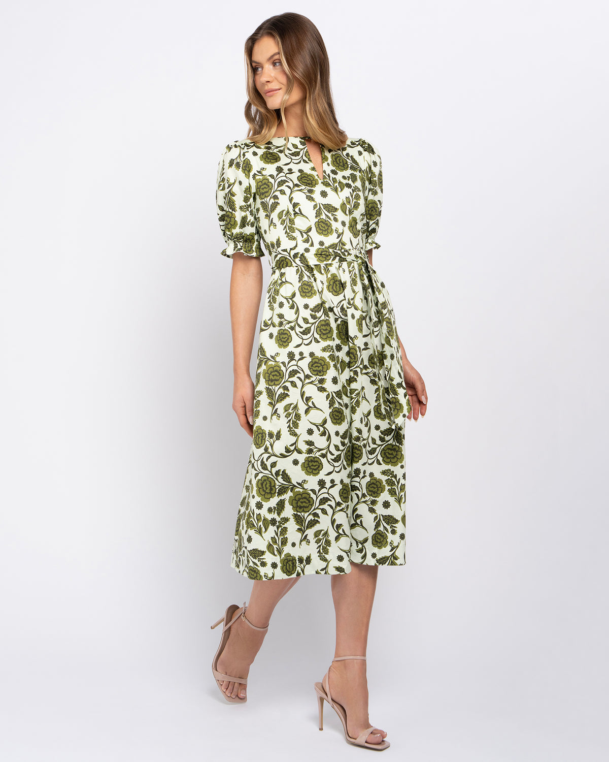 A woman modeling a knee-length dress with a green floral and botanical print on a white background, featuring puff sleeves, a keyhole neckline, and a self-tie waist belt, paired with beige high-heeled sandals against a light gray studio background.