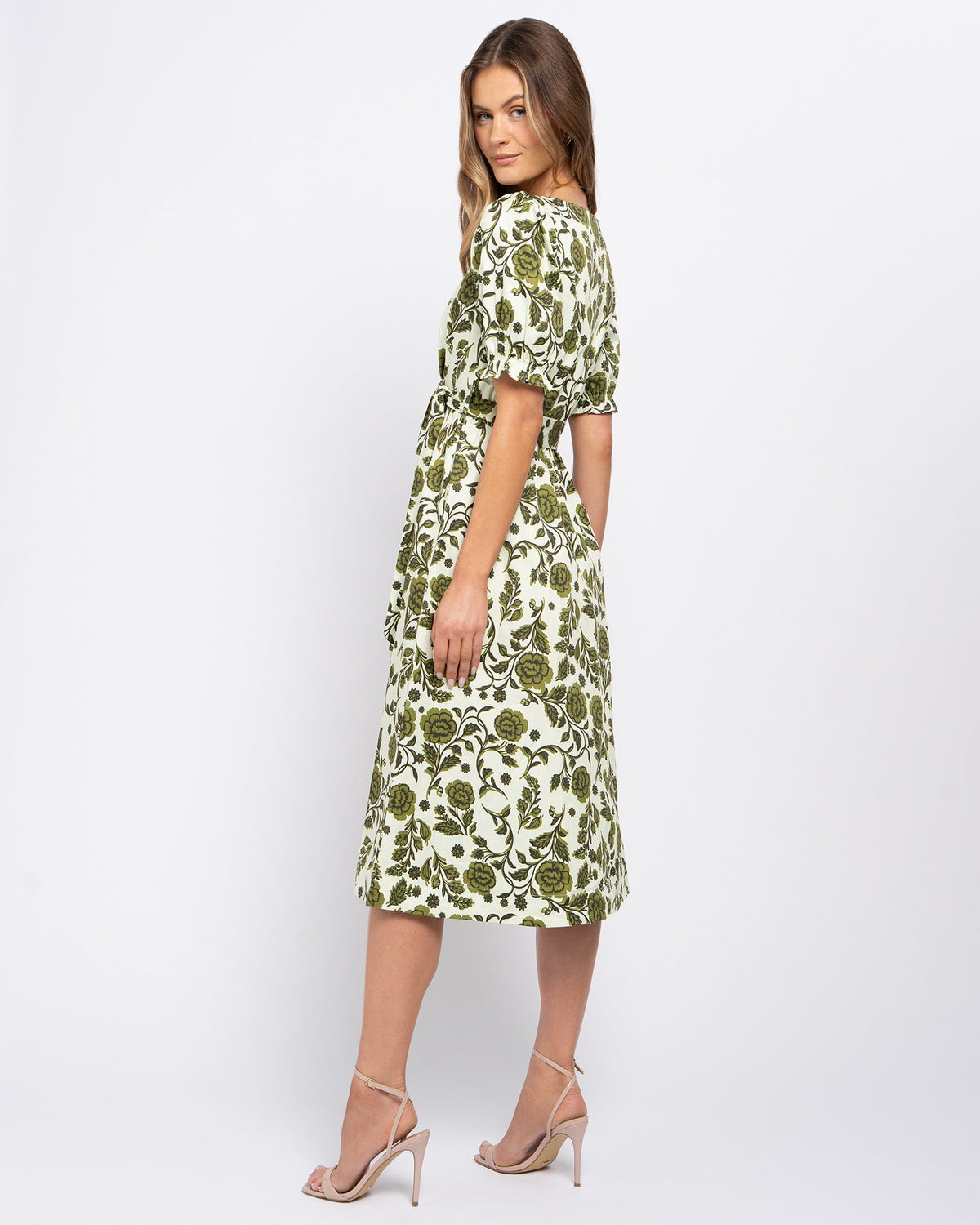 A woman modeling a knee-length dress with a green and white floral pattern, puff sleeves, and a tie at the back, shown from a three-quarter angle against a light gray studio background.