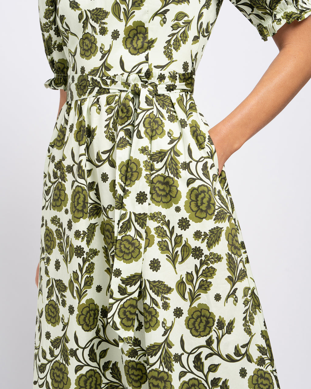 A close-up of a woman wearing a cream-colored dress with an olive green floral and vine pattern, featuring a tie waist and short flutter sleeves.