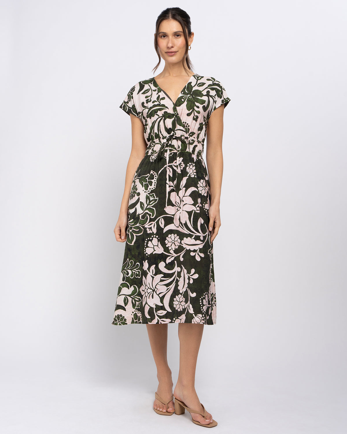 A woman modeling a knee-length wrap dress with a dark green and beige floral print, featuring short cap sleeves and a V-neckline, set against a plain light gray studio background.