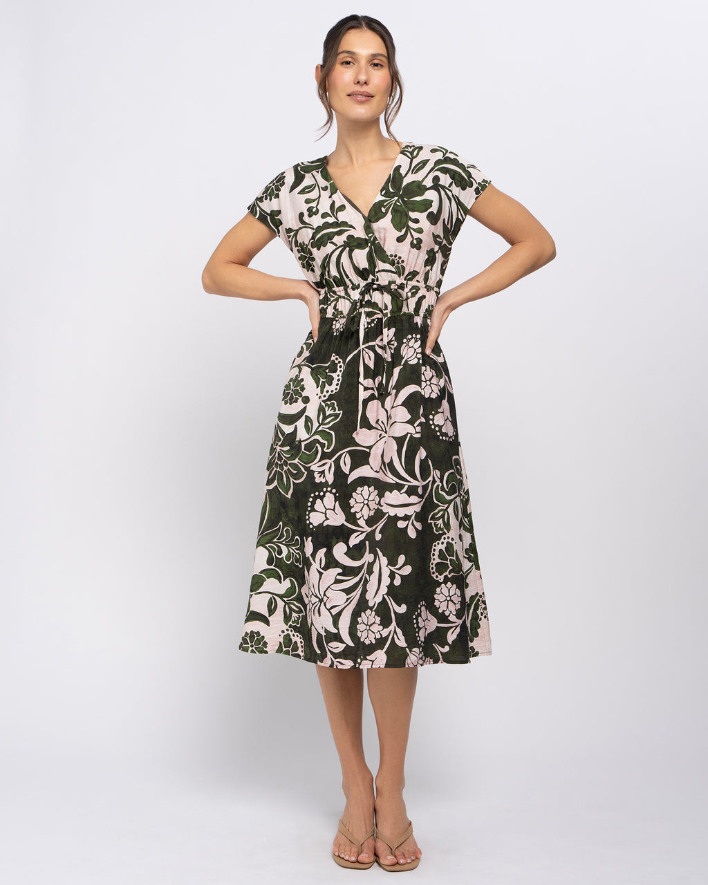 A woman modeling a short-sleeve midi dress with a dark green and beige floral pattern, standing against a light gray studio background.