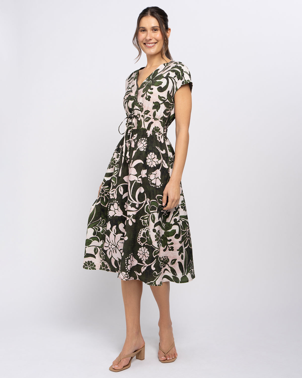 A woman modeling a knee-length wrap dress with a dark green, black, and light pink floral pattern, paired with beige heeled sandals, against a light gray studio background.