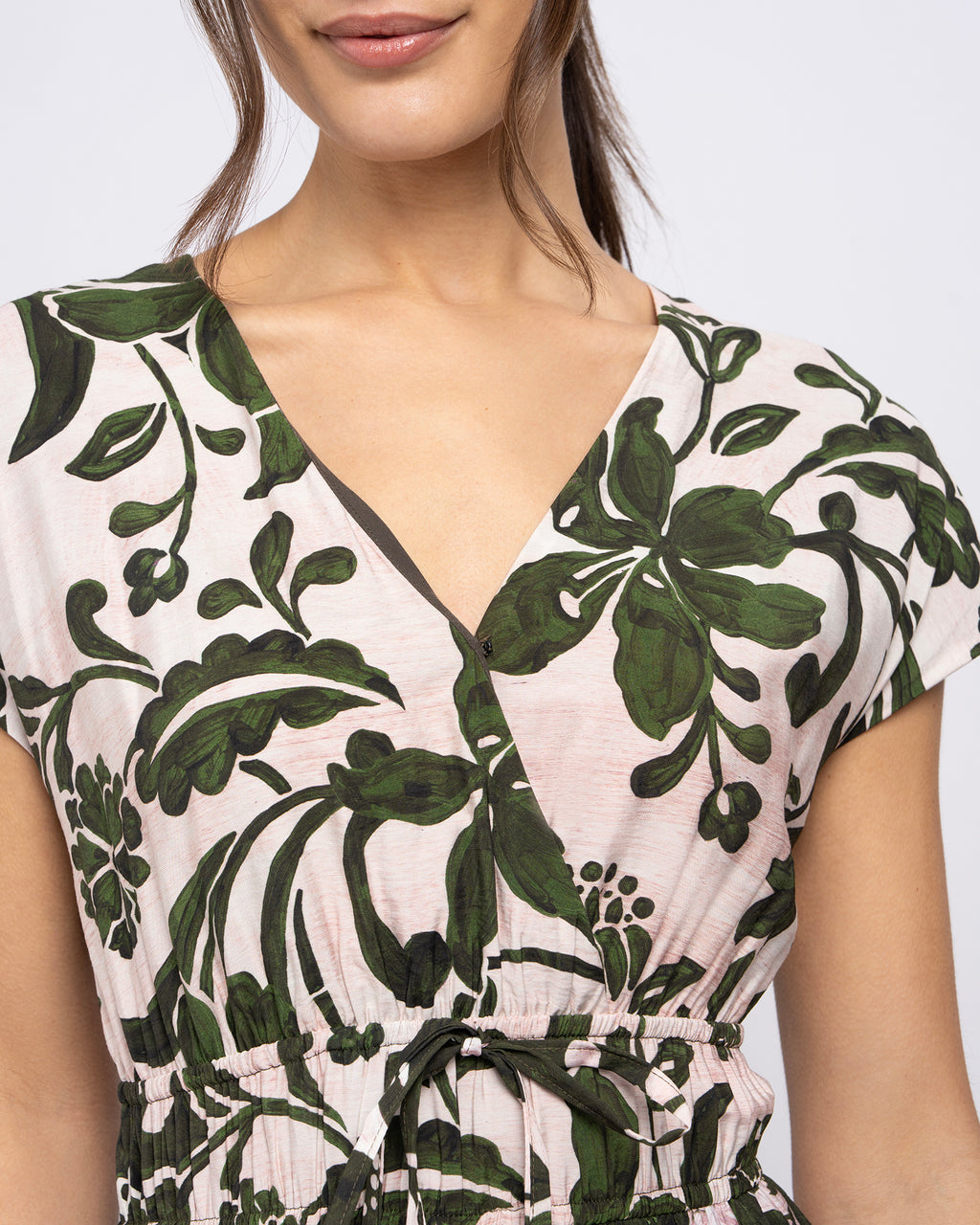 A close-up of a woman wearing a short-sleeve wrap-style top with a dark green floral print on a light pinkish-beige background.