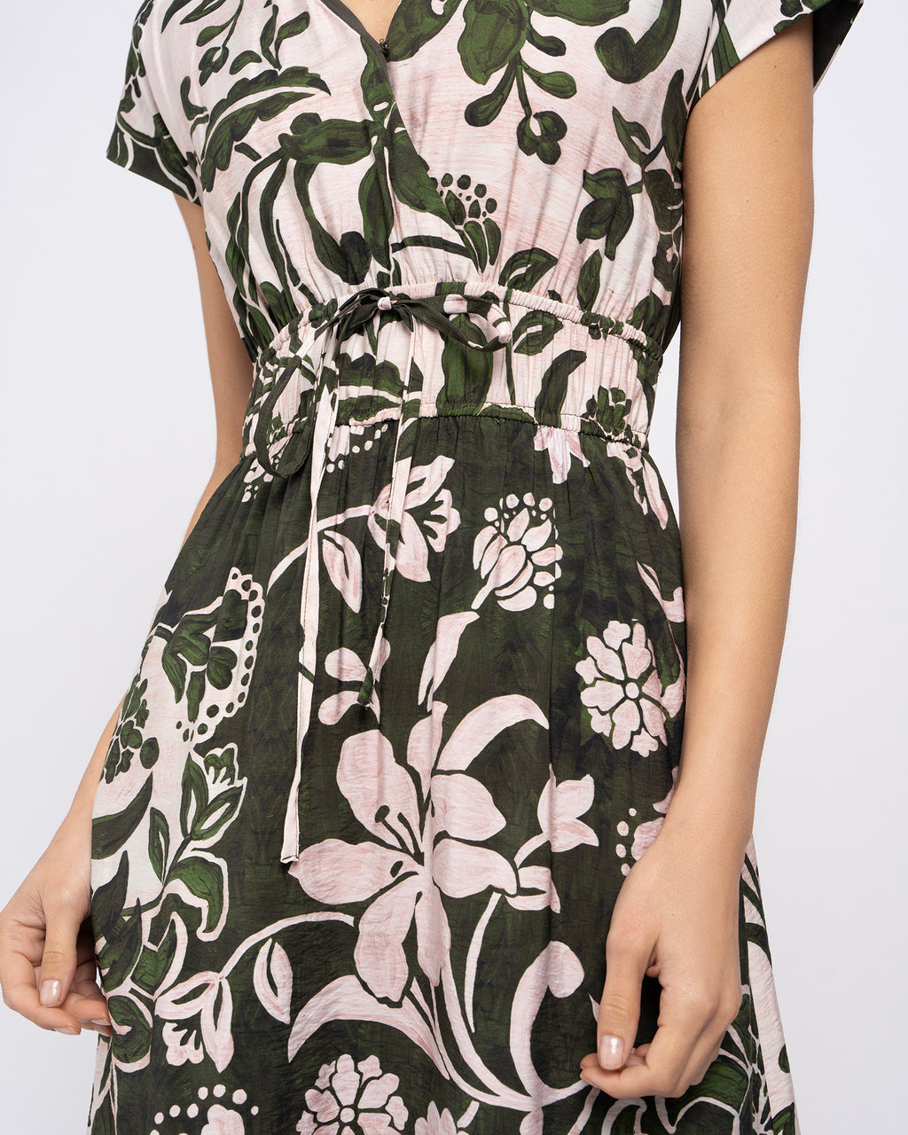 A close-up of a woman wearing a short-sleeve V-neck dress with a dark green, white, and pale pink floral print and a drawstring waist.