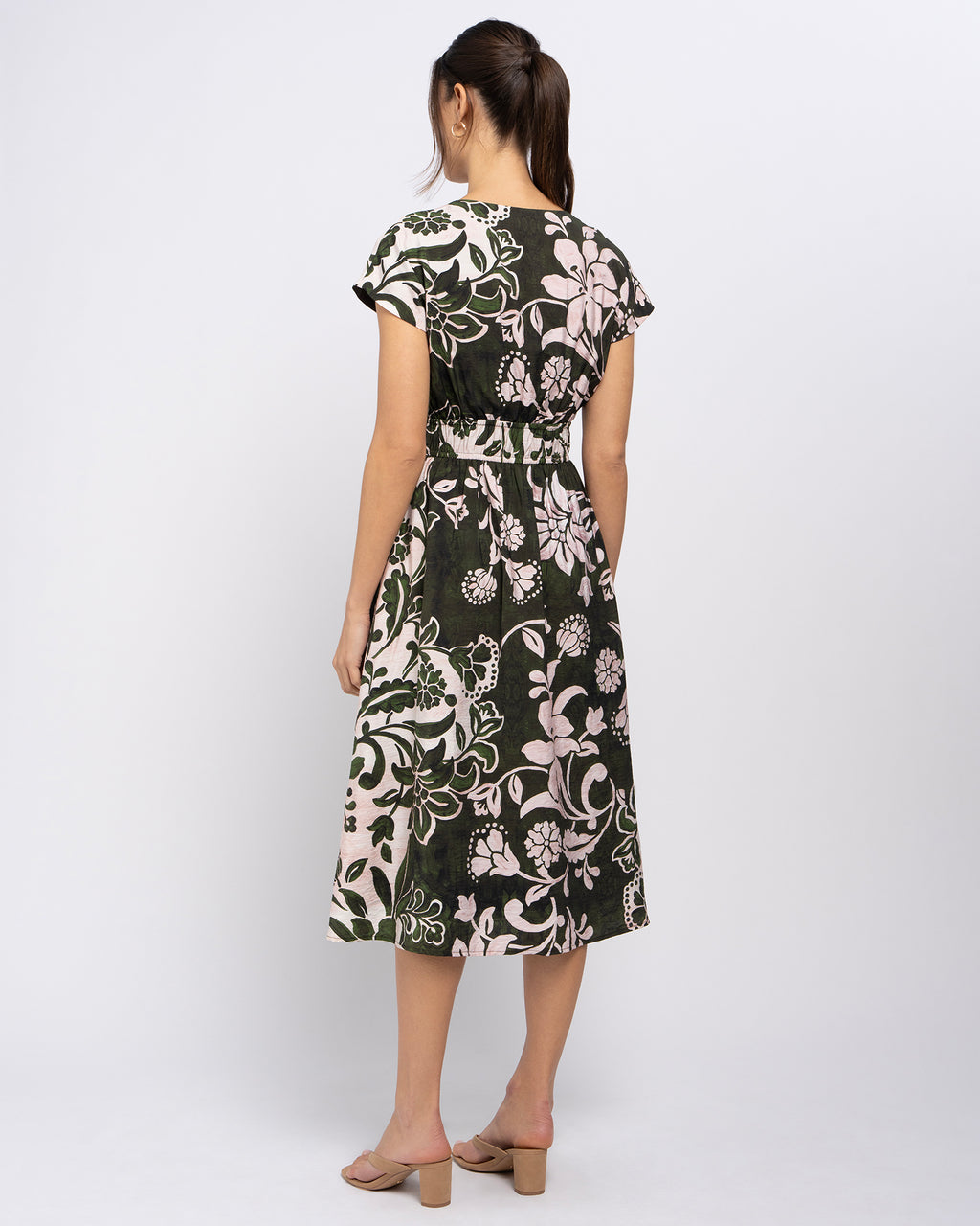 A woman viewed from the back wearing a knee-length dress with short cap sleeves and a floral print. The dress has a dark green base with large cream and light pink floral motifs, and a matching fabric tie at the waist.