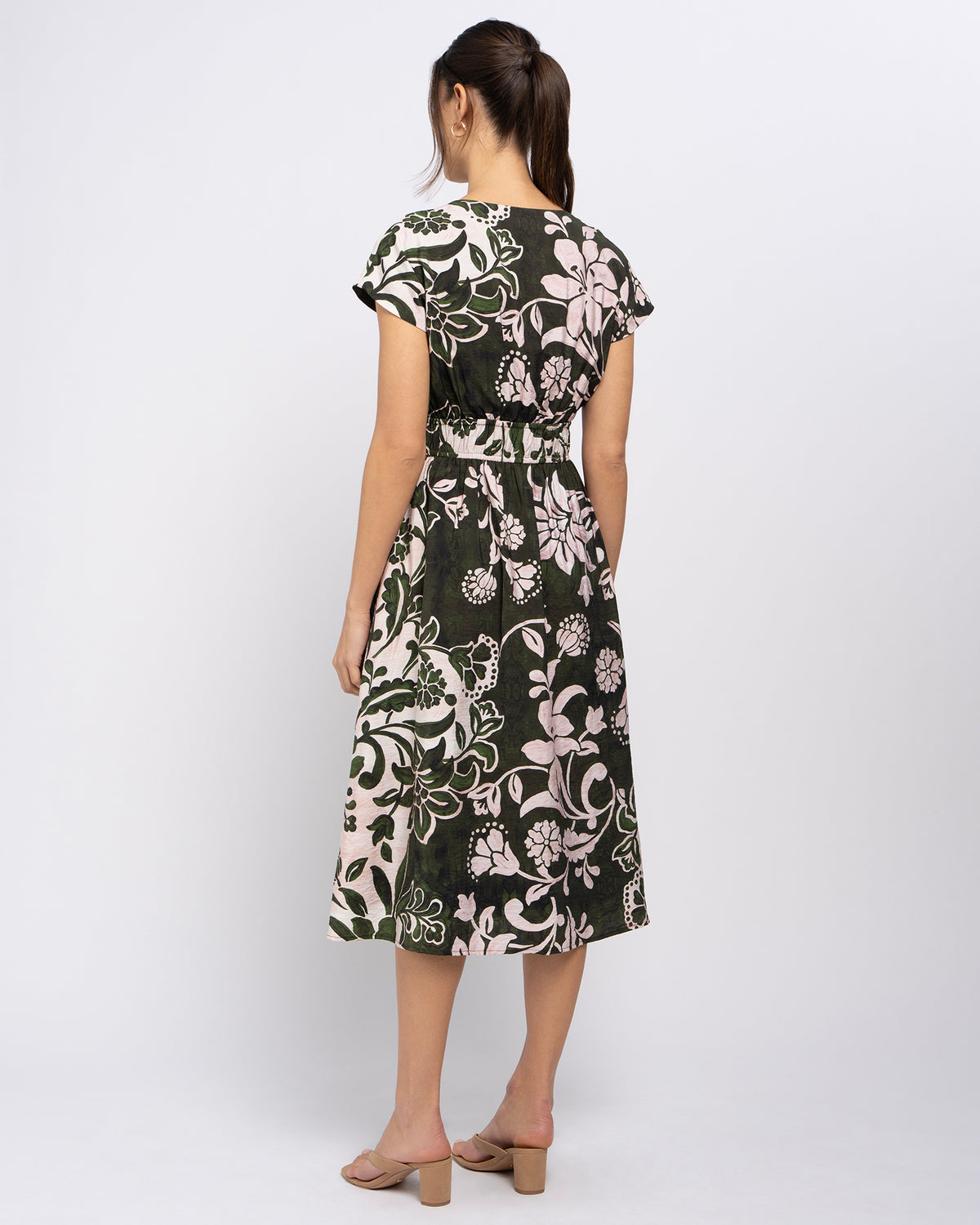 A woman viewed from the back wearing a knee-length dress with short cap sleeves and a floral print. The dress has a dark green base with large cream and light pink floral motifs, and a matching fabric tie at the waist.