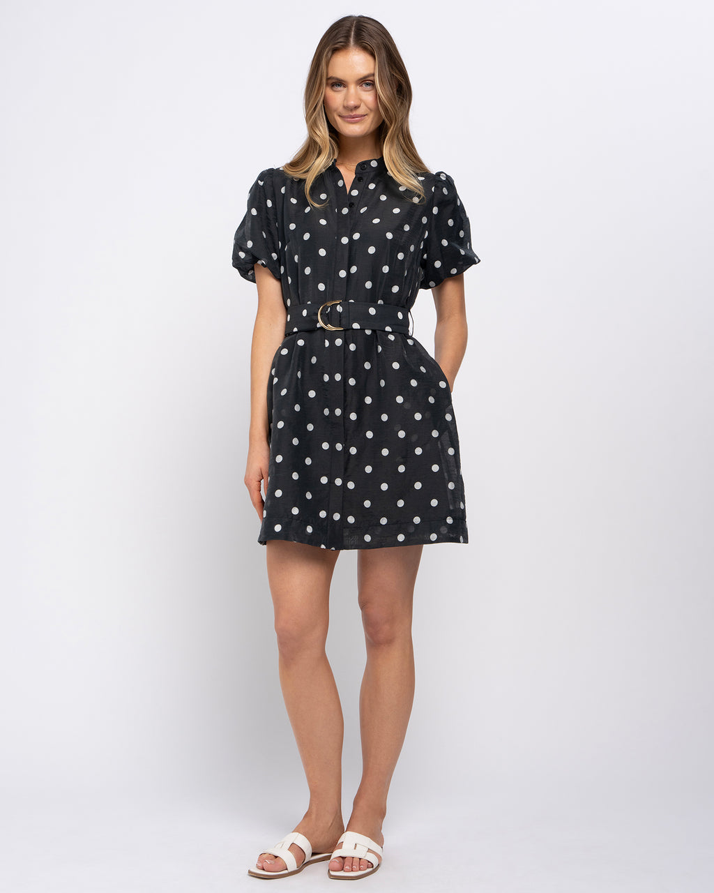 A woman modeling a black short-sleeve mini dress with white polka dots, featuring a pointed collar, puff sleeves, and a matching belt with a gold-tone buckle, set against a plain light gray studio background.