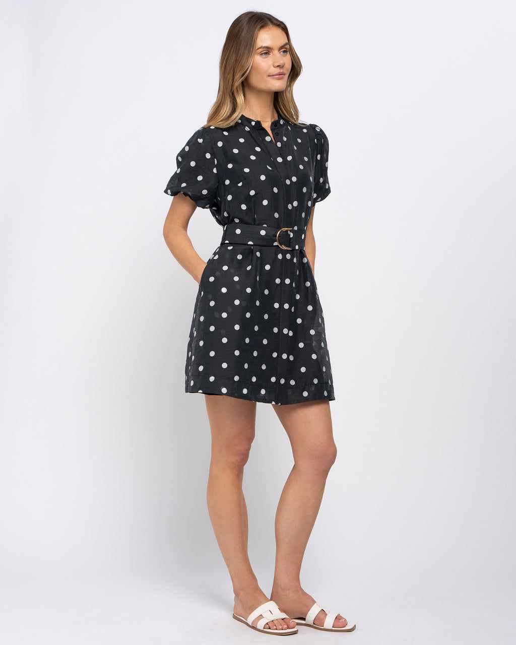 A woman modeling a short black dress with white polka dots, featuring puff sleeves, a pointed collar, and a matching waist belt with a gold-tone buckle, paired with white slide sandals against a light gray studio background.
