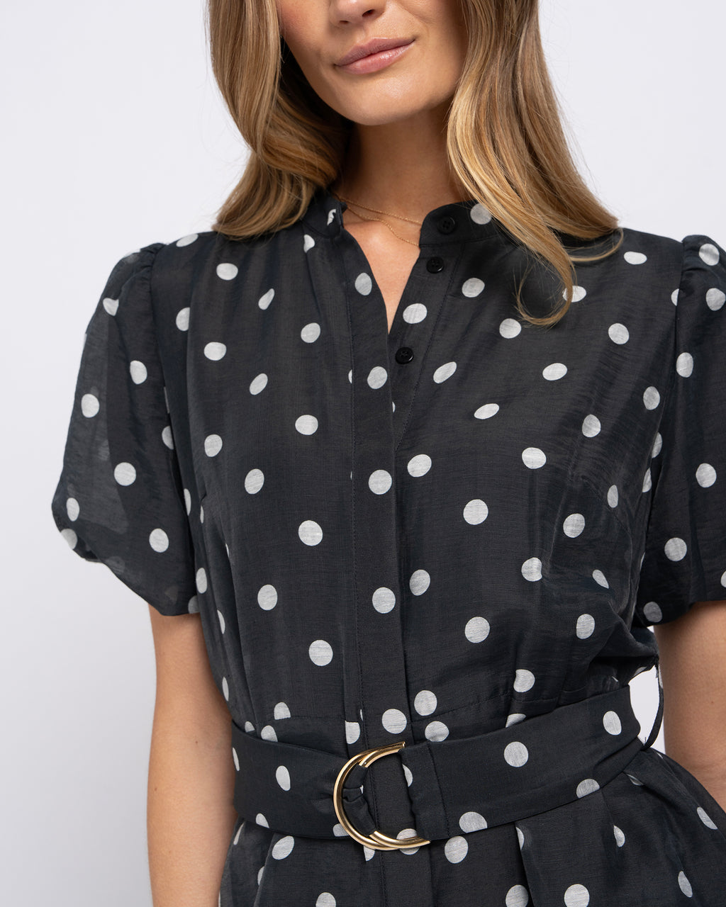 A close-up of a woman wearing a black short-sleeve blouse with white polka dots, featuring a button-down front, ruffled shoulders, and a matching waist belt with a gold-tone ring buckle.