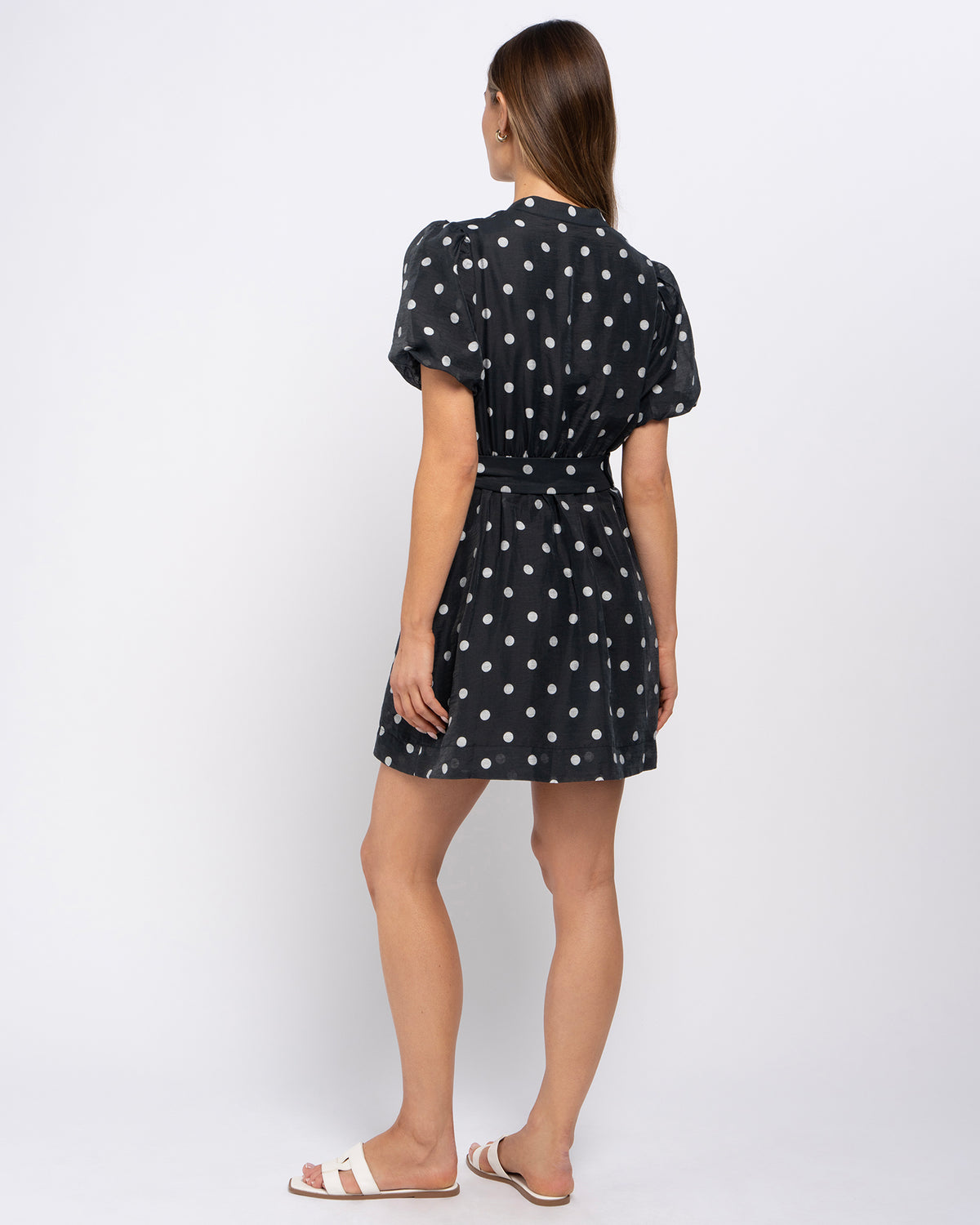 A woman modeling a short black dress with white polka dots, shown from the back against a plain white studio background.