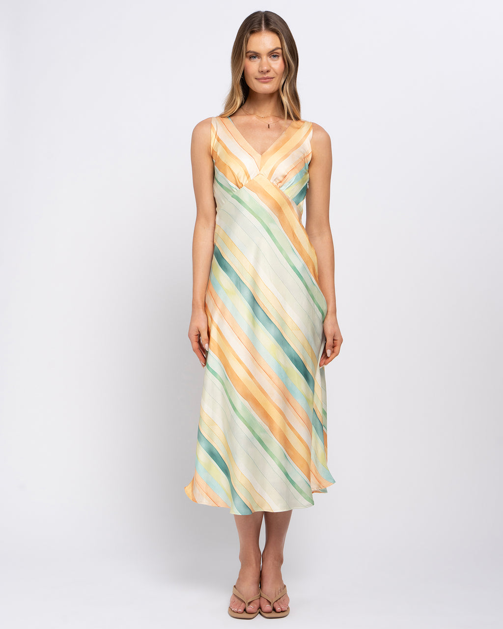 A woman modeling a sleeveless midi dress with a draped wrap-style bodice and diagonal striped pattern in pastel orange, teal green, and cream, set against a plain light gray studio background.