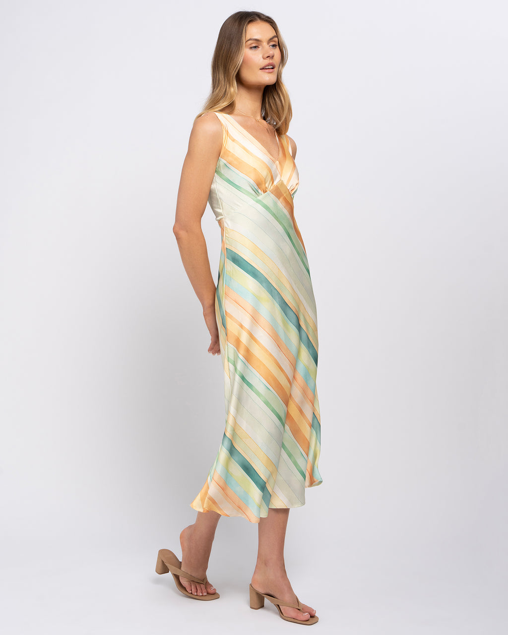 A woman modeling a sleeveless midi dress with diagonal stripes in cream, orange, teal, and yellow, paired with tan heeled sandals against a light gray studio background.