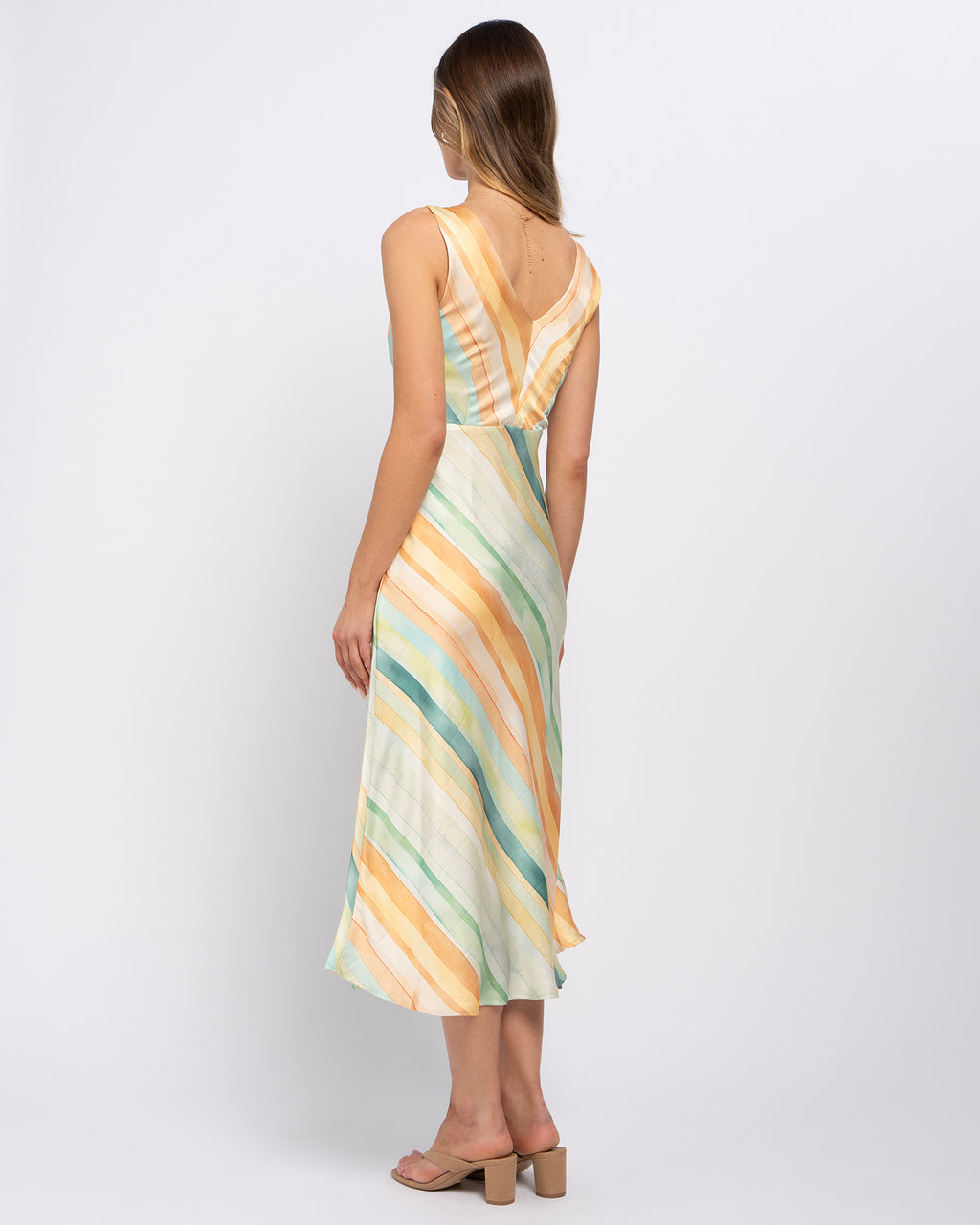 A woman shown from the back wearing a sleeveless midi dress with diagonal pastel stripes in yellow, orange, green, and cream, paired with beige block-heel sandals, against a light gray studio background.
