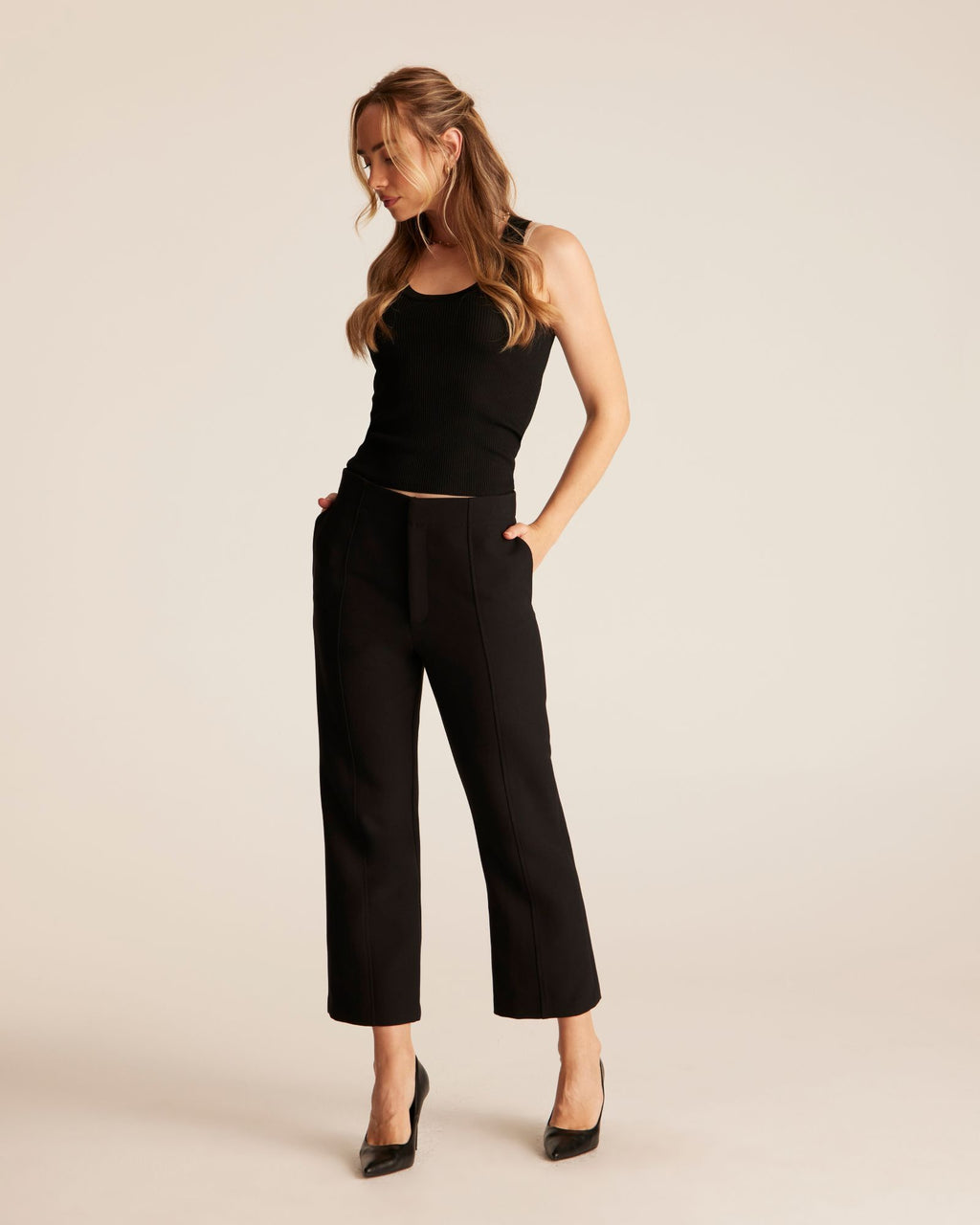 A woman modeling a black sleeveless crop top and black cropped wide-leg pants with black pointed heels, standing against a plain light beige studio background.