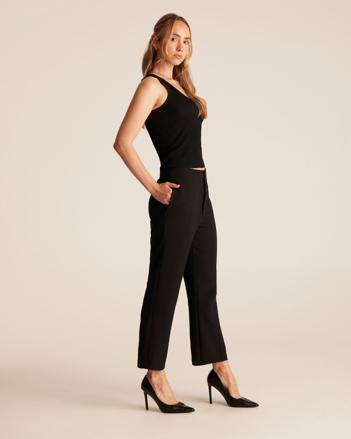 A woman modeling a black sleeveless top and matching black cropped trousers with black pointed-toe heels, posed against a plain warm beige studio background.