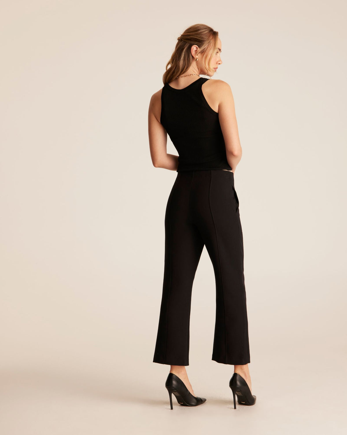A woman modeling a black sleeveless jumpsuit with high-waisted, wide-leg cropped pants, shown from the back against a solid light beige studio background.