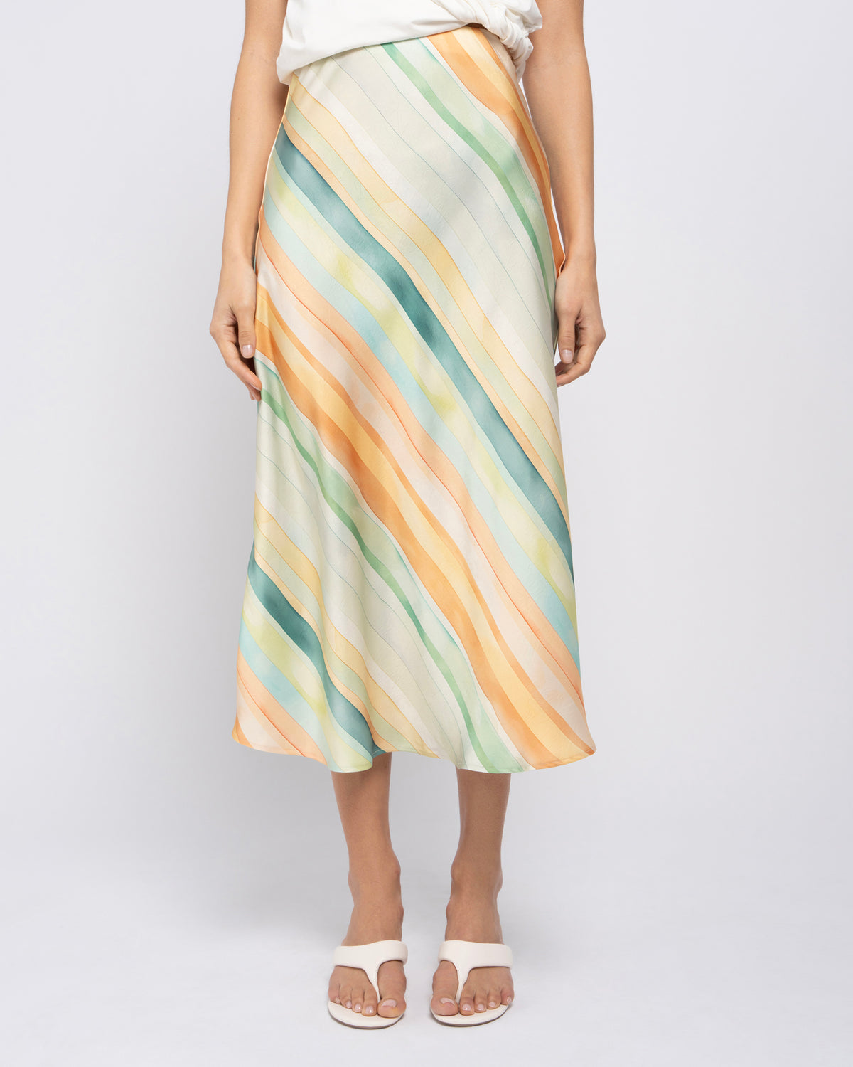 A model wearing a midi skirt with diagonal watercolor-style stripes in pastel yellow, mint green, and teal on a cream base, paired with white slide sandals against a light gray studio background.