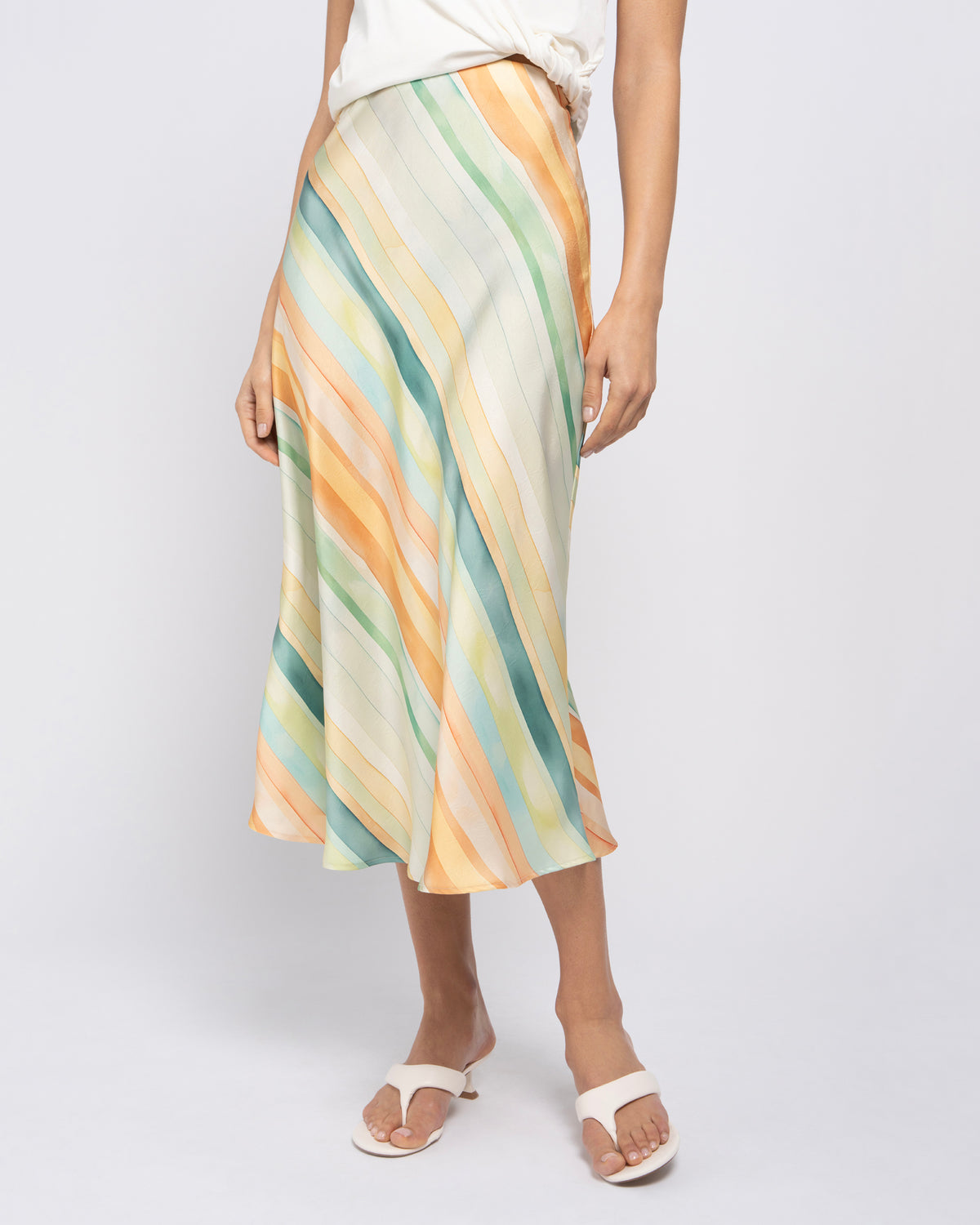 A model wearing a midi skirt with diagonal stripes in pastel orange, yellow, green, and blue on an off-white base, paired with a white top and white slide sandals, against a light gray studio background.