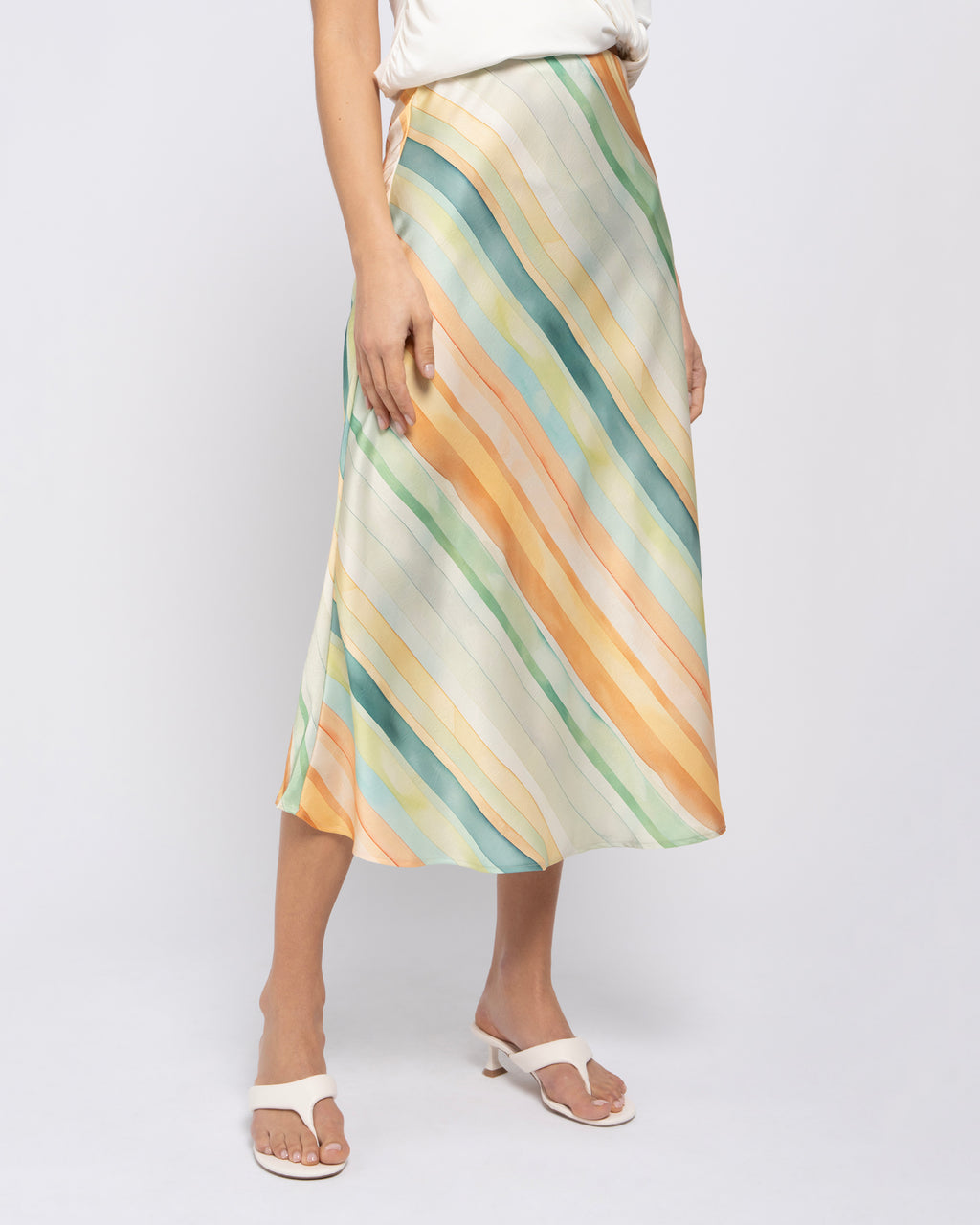 A model wearing a midi skirt with diagonal stripes in pastel shades of orange, green, blue, and cream, paired with white slide sandals against a light gray studio background.