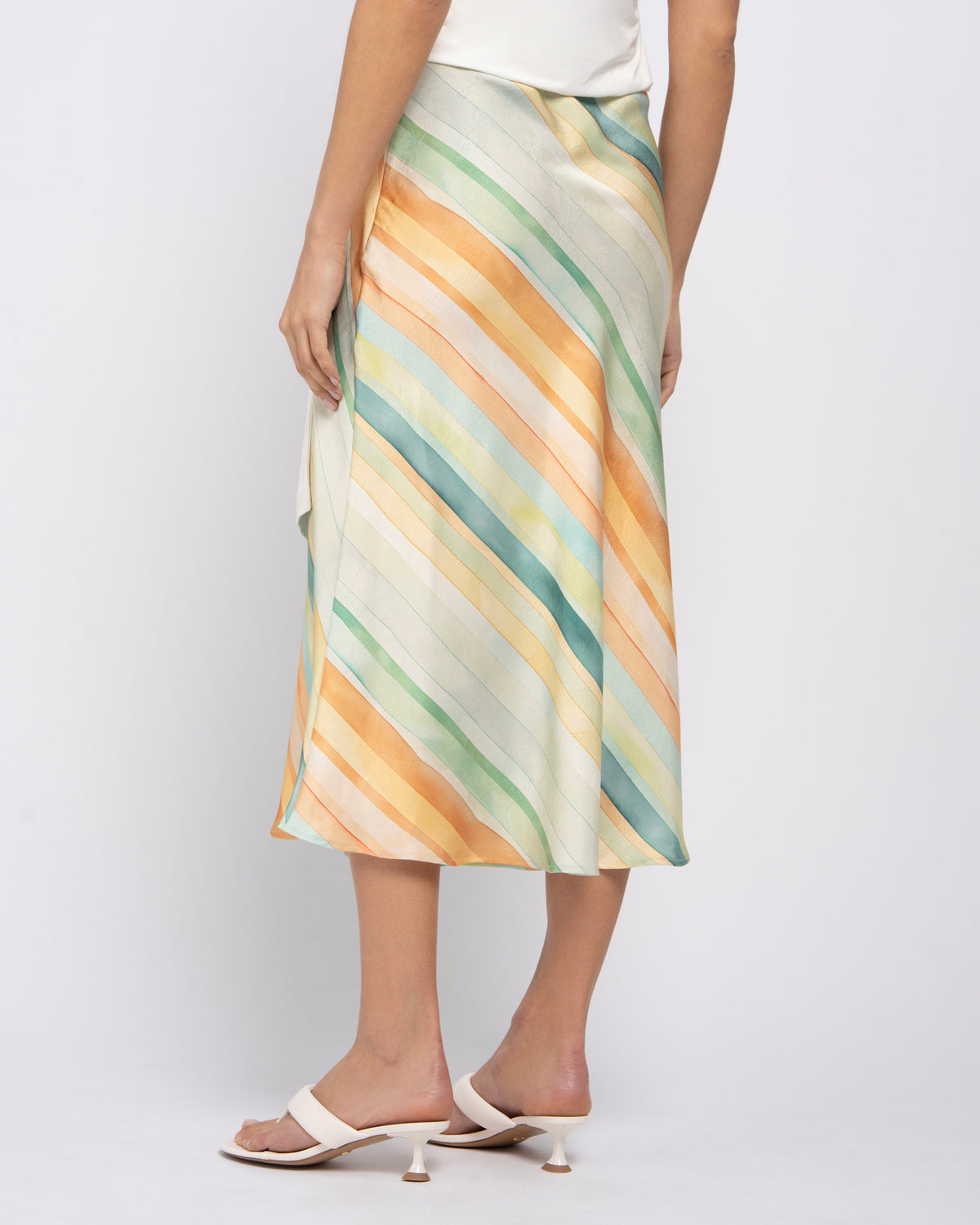 A model is shown from the waist down wearing a midi skirt with diagonal stripes in pastel shades of yellow, orange, green, and blue on a cream base. The model is also wearing white slide sandals against a plain light gray studio background.