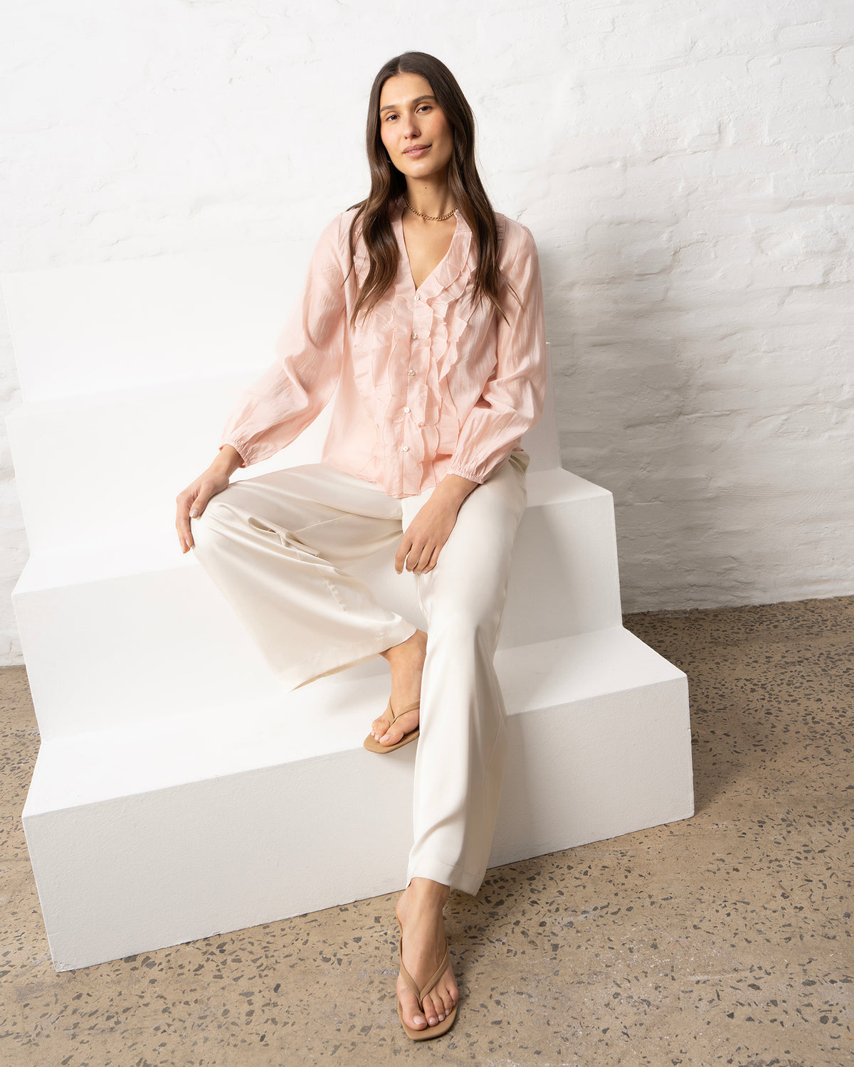 A woman sits on white geometric steps against a white brick wall, wearing a light blush pink ruffled blouse and cream wide-leg trousers.