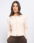 Stella Women's Beatrice Knit - Ballerina. A woman wearing a light beige long-sleeve button-up cardigan and dark brown pants, photographed against a plain light gray studio background.