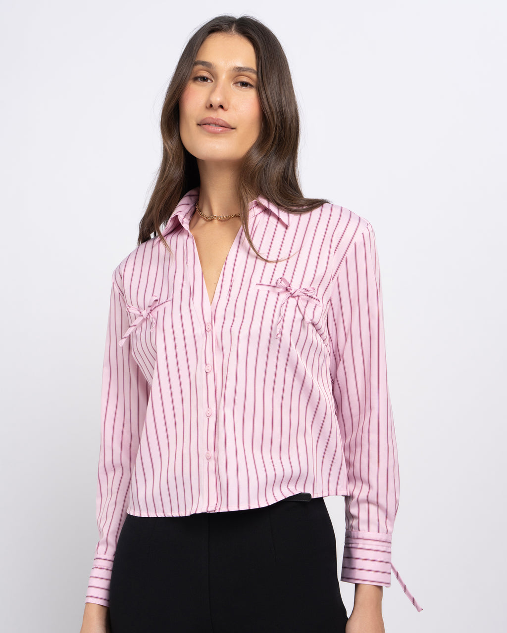Stella Women's Jenny Shirt - Pink Candy Stripe. A woman wearing a pink and white vertically striped button-up blouse with ruffled chest details and tie cuffs, paired with black pants, against a light gray studio background.