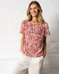 Stella Women's Margot Top - Pink Ditsy. A woman wearing a dusty rose pink short-sleeve blouse with a white floral pattern, paired with cream-colored trousers, standing against a white brick wall.